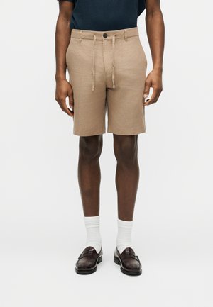 Homme portant un short beige avec cordon de serrage, des chaussettes blanches et des mocassins marron foncé, debout devant un fond uni.