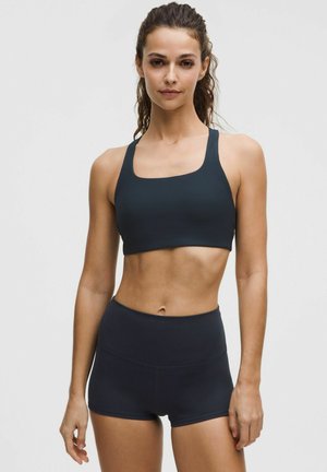 Mujer con cabello largo vestido con un sujetador deportivo oscuro y pantalones cortos a juego, de pie mirando hacia adelante sobre un fondo claro liso.