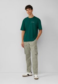 Jongeman staat met zijn handen in zijn zakken, gekleed in een groen T-shirt, beige cargo-broeken en witte sneakers met zwarte accenten.