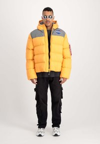 Chaqueta acolchada de color amarillo brillante con hombros grises, que presenta un parche de la NASA, cierre de cremallera y un diseño corto. Se lleva con pantalones cargo negros.