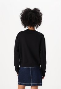 Schwarzer Pullover mit einem gerippten Kragen und Bündchen. Getragen mit einem Jeansrock, der Nähte und einen Knopfakzent hat. Einfarbiger Hintergrund.