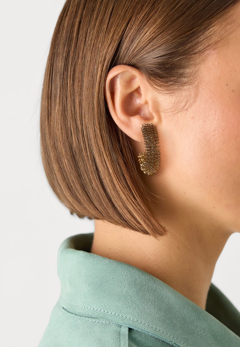 Goldfarbene, strukturierte Ohrringe mit einer einzigartigen Form, die ein mehrlagiges Design und filigrane Details aufweisen, die an dem Ohr eines Models getragen werden.