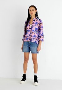 Camicia floreale viola a maniche corte abbinata a pantaloni corti in denim. Il modello indossa sneaker alte nere con suola bianca, su uno sfondo bianco.