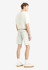 Pantalones cortos de mezclilla verde claro con un ajuste relajado, un parche de cuero en la parte posterior y bolsillos traseros cosidos. Llevados con una camiseta blanca y zapatillas.