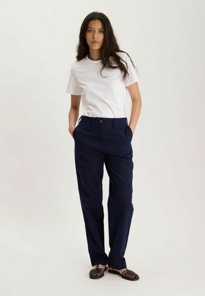 Jeune femme aux longs cheveux foncés portant un t-shirt blanc à manches courtes, un pantalon bleu marine et des sandales tressées foncées, debout les mains dans les poches.