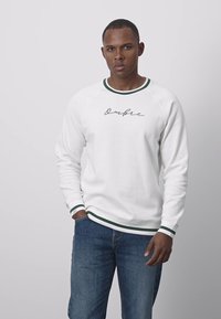 Vit sweatshirt med gröna randiga ribbade muddar och fåll. Har en text i skrivstil "Ombre" på framsidan. Kombinerad med blå jeans.