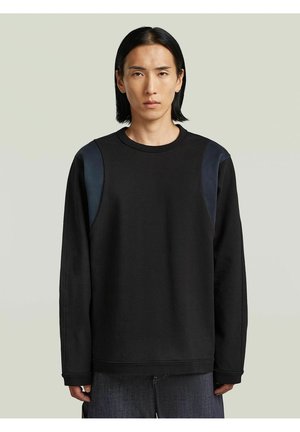 Giovane uomo con capelli neri lisci che indossa una camicia nera a maniche lunghe con toppe scure sulle spalle e pantaloni scuri su uno sfondo semplice.