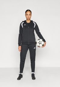 Μαύρη φόρμα Adidas με φερμουάρ στη κουκούλα, λευκές λεπτομέρειες και παντελόνι που ταιριάζει. Μαύρα αθλητικά παπούτσια. Κρατάει μια ποδοσφαιρική μπάλα με σχέδια.