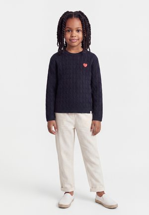 Enfant aux cheveux tressés portant un pull bleu marine avec un écusson en forme de cœur, un pantalon couleur crème et des chaussures blanches sans lacets, debout devant un fond uni.