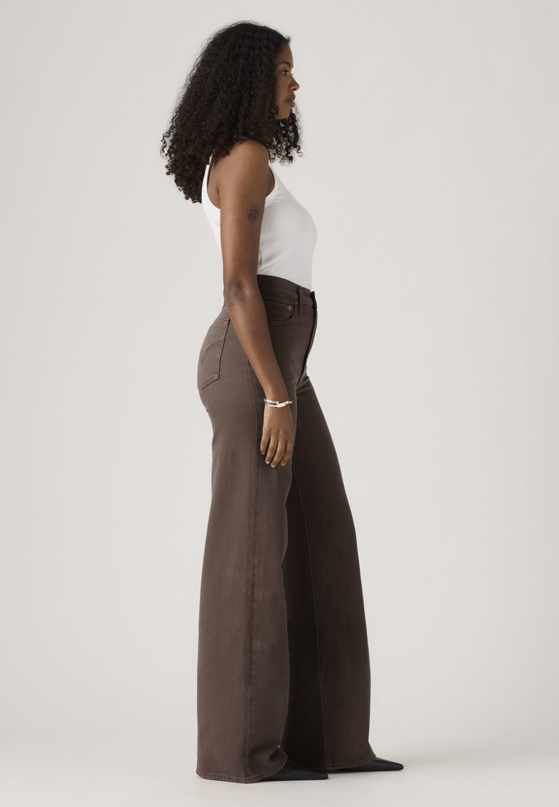 Femme aux cheveux bouclés portant un haut blanc sans manches et un pantalon marron taille haute à jambes larges, debout de profil devant un fond uni.