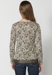 Langärmliges Shirt in Beige mit schwarzem Blumenmuster. Weicher Stoff, runder Halsausschnitt und strukturierter Saum. Wird mit dunklen Jeans getragen.