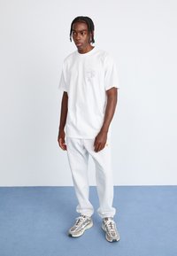 White cotton t-shirt with a small graphic on the left chest, paired with light gray sweatpants and neutral-toned athletic shoes.