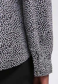 Poignet de chemise à manches longues à motif noir et blanc avec bouton, portée avec un pantalon noir, montrée en gros plan sur le bras d'une personne.
