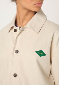 Beige Segeltuchjacke mit einem grünem, diamantförmigen Aufnäher mit gelbem Text. Verfügt über einen Kragen und zwei Knopfverschlüsse.