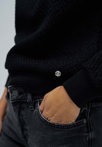 Černý svetr s texturou, ribbovanými manžetami a lemem, s malým stříbrným logem na boku. Nesen s tmavě šedými džínami.