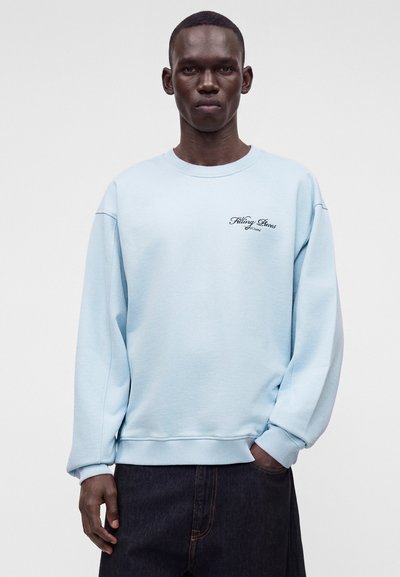 Homme portant un sweat-shirt bleu clair avec le texte "Filling Pieces" sur la poitrine et un jean foncé, debout devant un fond clair uni.