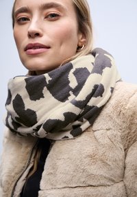 Écharpe infinie beige et grise à motifs, en tissu doux, enroulée autour du cou, associée à un manteau en fausse fourrure crème.