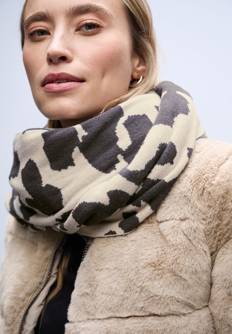 Écharpe infinie beige et grise à motifs, en tissu doux, enroulée autour du cou, associée à un manteau en fausse fourrure crème.