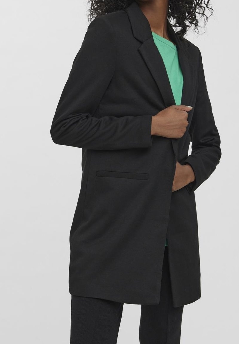 Personne portant un long blazer noir par-dessus une chemise verte, une main ajustant le blazer, l'autre dans la poche.