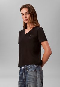 T-shirt nera a scollo a V in cotone, con un piccolo logo sul petto a sinistra. Maniche corte e taglio comodo. Indossata con jeans blu in denim.