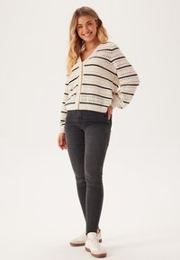 Strickjacke in Creme mit schwarzen Streifen und Lochmustern, kombiniert mit dunklen Jeans und weißen Sneakers. Lässig geschnitten und mit V-Ausschnitt.