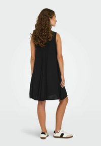 Vestido negro sin mangas con un diseño acampanado, a la altura de la rodilla, hecho de tejido ligero. Combinado con zapatillas blancas con detalles en negro.
