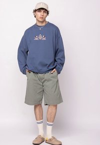 Sudadera azul extragrande con estampado gráfico, pantalones cortos grises, gorra beige y zapatos sin cordones de color beige claro. Calcetines blancos con rayas azules y amarillas.