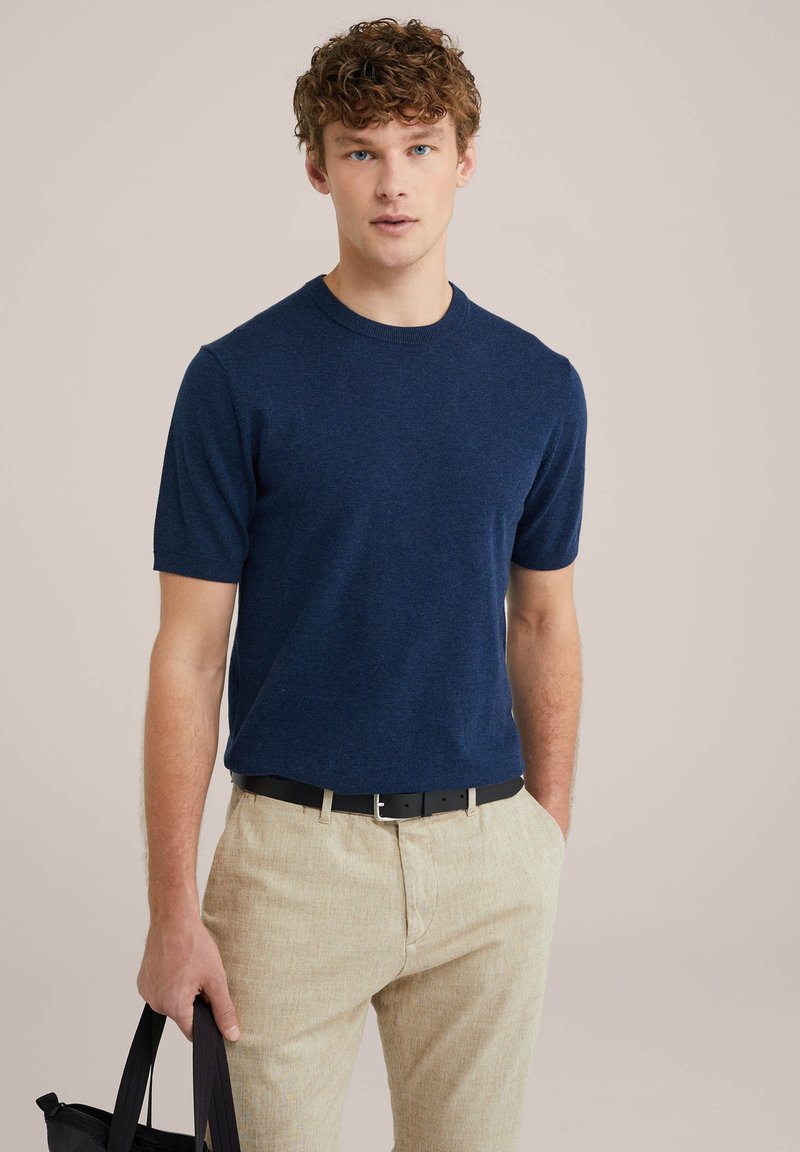 Tamsiai mėlynos spalvos trumpomis rankovėmis crew neck marškiniai, minkšta tekstūra, dera su šviesiai smėlio spalvos tekstūruotomis kelnėmis ir juodu diržu, laikomais juodu krepšiu.