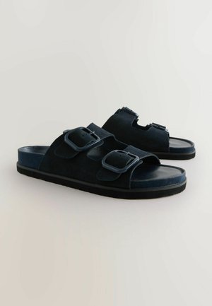 Tume sinine sametist libisemisjalanõud, millel on kaks reguleeritavat rihma, millel on ristkülikukujulised pandlad, ja lame kummist tõstepõhi. Sile sisekeel.