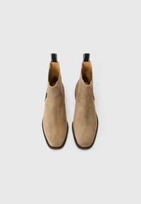 Bottines Chelsea en suède beige avec un bout arrondi, des panneaux élastiques sur les côtés et une languette noire à l'arrière ; semelle plate en caoutchouc noir.