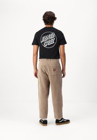 Svart t-shirt med vitt cirkulärt "Santa Cruz"-logotyp, matchad med beige kordbyxor och svarta skor, sedd från baksidan.