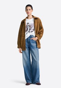 Brūna zamša jaka virs baltas grafiskās T-kreklas; vaļīgas zilas džinsu bikses; bordo kurpes; ikdienišķs, brīvs piegriezums; modelis stāv pret vienkāršu fonu.