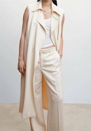 Longue veste beige avec un col cranté, portée sur un débardeur blanc et un pantalon beige taille haute. Tissu lisse et coupe ample.