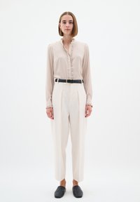 Camicia button-up color beige chiaro con colletto arricciato, abbinata a pantaloni larghi crema e scarpe nere slip-on; design minimalista.