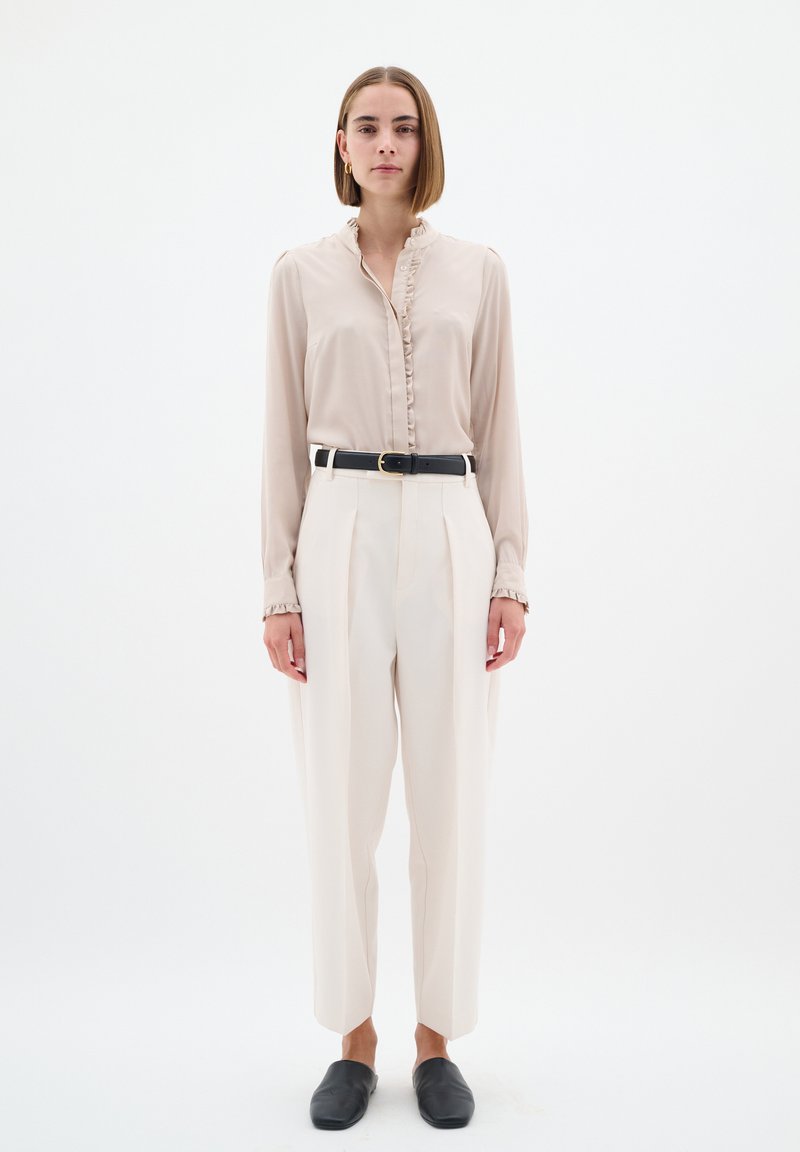 Camicia button-up color beige chiaro con colletto arricciato, abbinata a pantaloni larghi crema e scarpe nere slip-on; design minimalista.