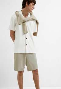 Hvit kortermet skjorte med knapper og fin tekstur, kombinert med lysebeige shorts. En beige strikkegenser henger over skuldrene.