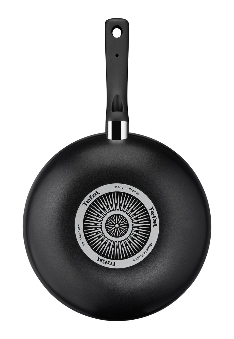 Zwarte koekenpan met een lange handgreep, cirkelvormig patroon aan de onderkant, en "Tefal" branding, gezien vanaf de onderkant op een witte achtergrond.