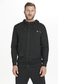 Hoodie noir zippé fabriqué dans un tissu lisse, avec un logo sur la poitrine et une capuche avec des cordons de serrage. Il comprend des poches avant.