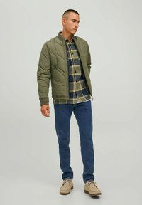 Chaqueta acolchada oliva con cuello alto, superpuesta a una camisa de cuadros en azul y beige, combinada con jeans azules y zapatos beige.