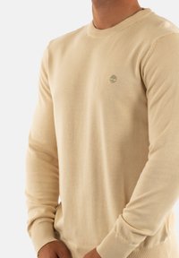 Maglione beige a maglia con polsini e orlo a coste. Presenta un piccolo logo di un albero verde sul petto. Il materiale sembra morbido e liscio.