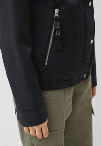 Veste noire légère avec une poche zippée, des boutons à pression et une taille élastique. Présente une texture lisse et un design décontracté.