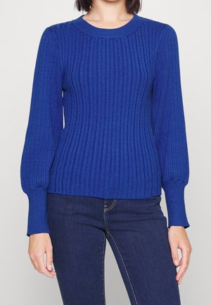 Persona que lleva un suéter ajustado de manga larga color azul real con textura de canalé, combinado con vaqueros azul oscuro, de pie frente a un fondo blanco liso.