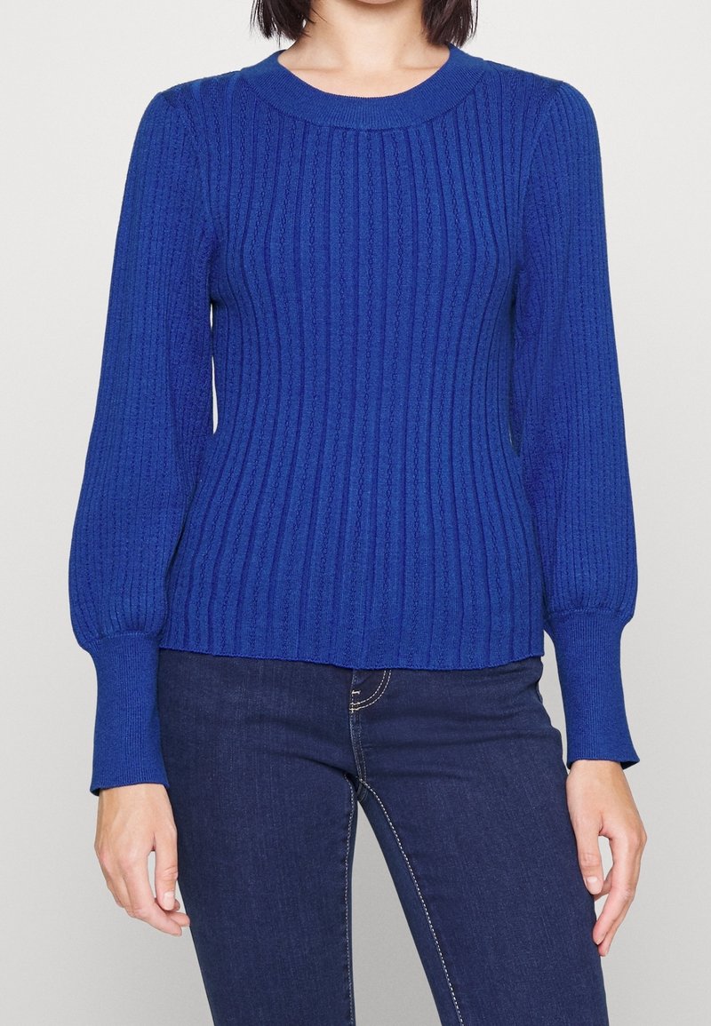 Personne portant un pull ajusté à manches longues bleu roi en maille côtelée, associé à un jean bleu foncé, debout devant un fond blanc uni.