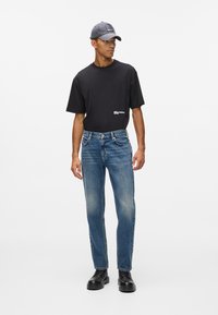 Juodas marškinėliai trumpomisRankovėmis su logotipu, mėlyni džinsai ir juodi auliniai batai su raišteliais. Modelis dėvi pilką kepurę, demonstruodamas kasdienį aprangą.