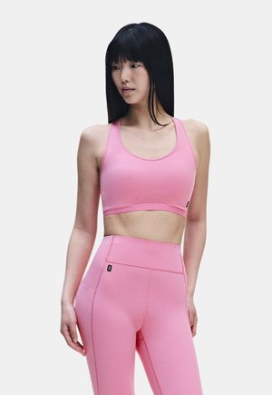 Frau mit langen schwarzen Haaren, die einen pinkfarbenen Sport-BH und hoch taillierte pinke Leggings trägt und nach links schaut vor einem einfarbigen hellen Hintergrund.