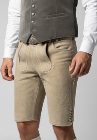 Krémové kožené šortky s dekorativní výšivkou, bočními kapsami a knoflíkovými detaily na lemu. V kombinaci s šedou vestou a bílou košilí.