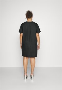 Fekete ruha rövid fodros ujjakkal, egyenes szabással és térdhez érő hosszúsággal. Ezüst magas sarkú cipővel viselve.