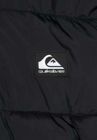 Melna pūkainā jakā ar gludu, vieglu audumu, uz krūtīm izvietota balta Quiksilver logo uzšuve un horizontālās šuves detaļa.