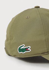 Oliven grøn caps med en teksturert overflate og buet skjerm, med en hvit lapp med en grønn krokodillelogo og røde detaljer.