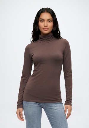 Modèle portant un col roulé marron ajusté à manches longues et un jean bleu clair sur un fond blanc uni.